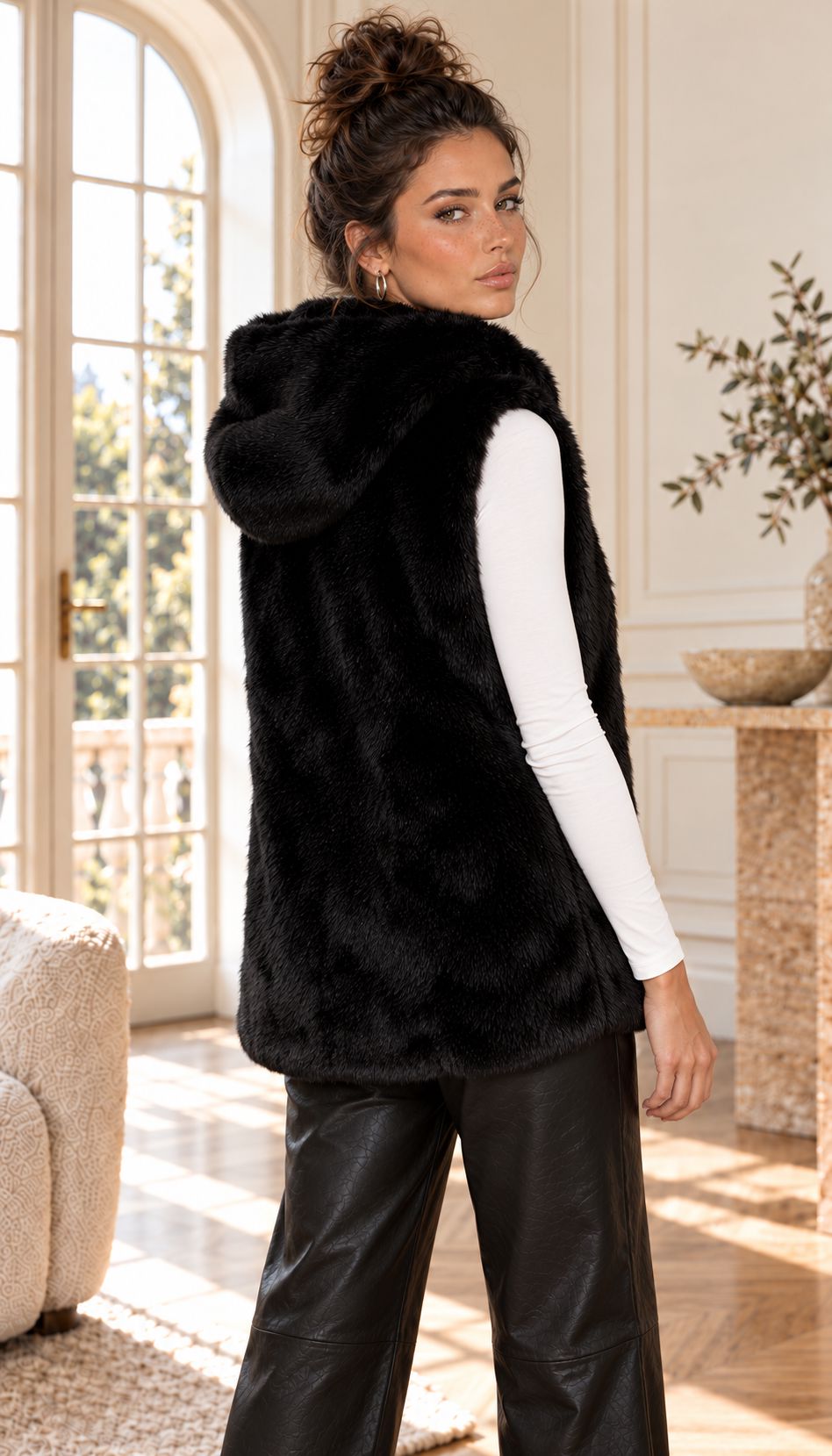A woman models a black faux fur zip-up vest with a plush collar, worn over a white long-sleeve top and black leather pants in a bright, elegant room.