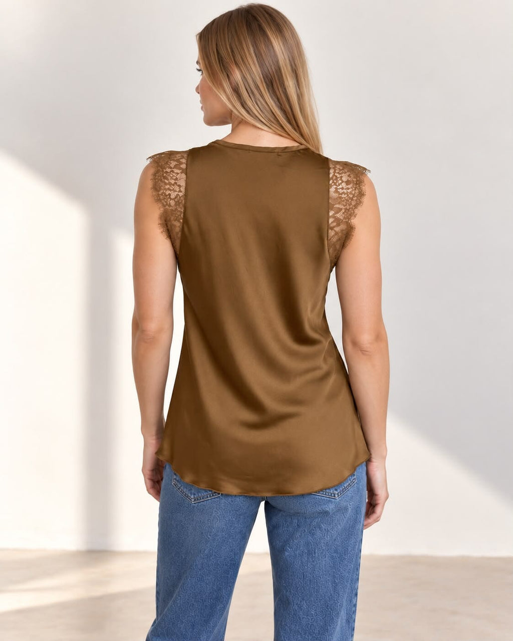 A woman modeling a sleeveless bronze satin top with a lace V-neckline, paired with blue jeans, standing against a neutral studio wall.