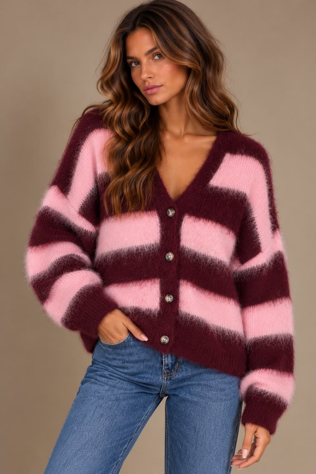 A woman models a fuzzy knit cardigan with horizontal stripes in dusty pink and deep burgundy, paired with wide-leg blue jeans and burgundy flats, set against a warm beige studio background with soft window shadows.
