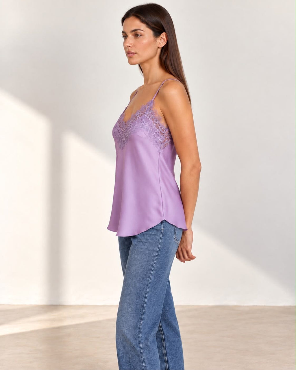 A woman modeling a lavender satin camisole top with floral lace cups and thin spaghetti straps, paired with blue jeans, in a brightly lit studio setting.