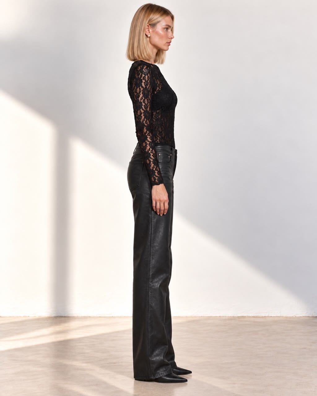 A model stands in a minimalist studio wearing a black lace long-sleeve top and black leather wide-leg pants.