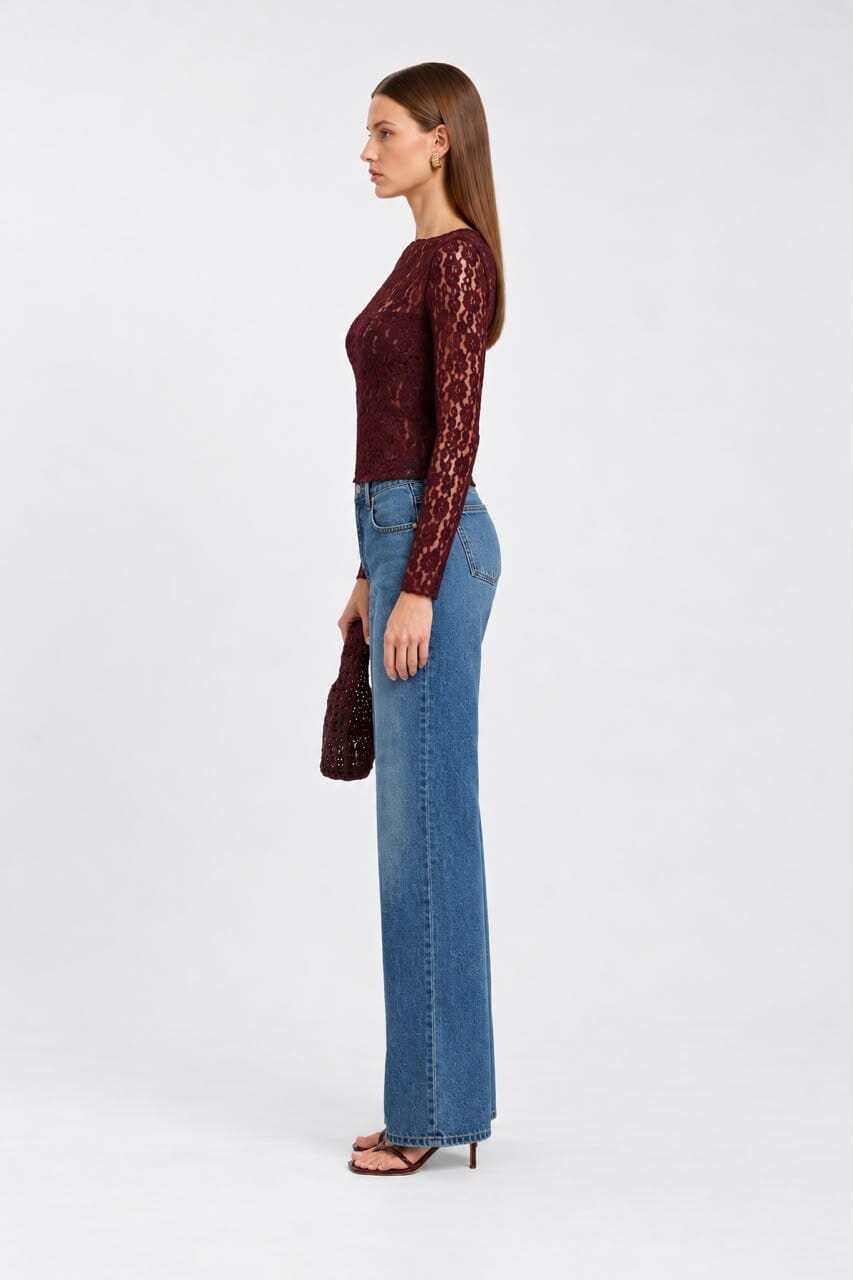 A model stands against a light gray studio background wearing a long-sleeve burgundy lace top, blue flare jeans, and black sandals, while holding a small matching burgundy clutch.