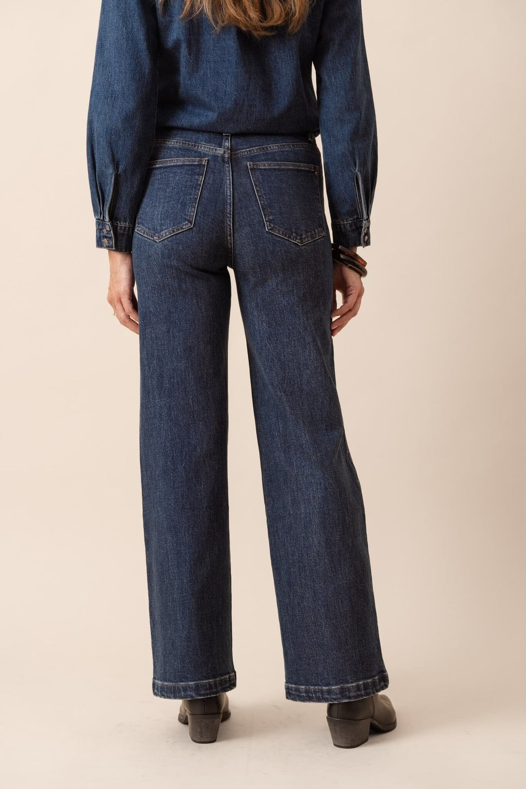 A woman modeling a dark blue denim long-sleeve button-up shirt with a tie-neck detail and matching dark blue denim jeans, standing against a solid beige studio background.